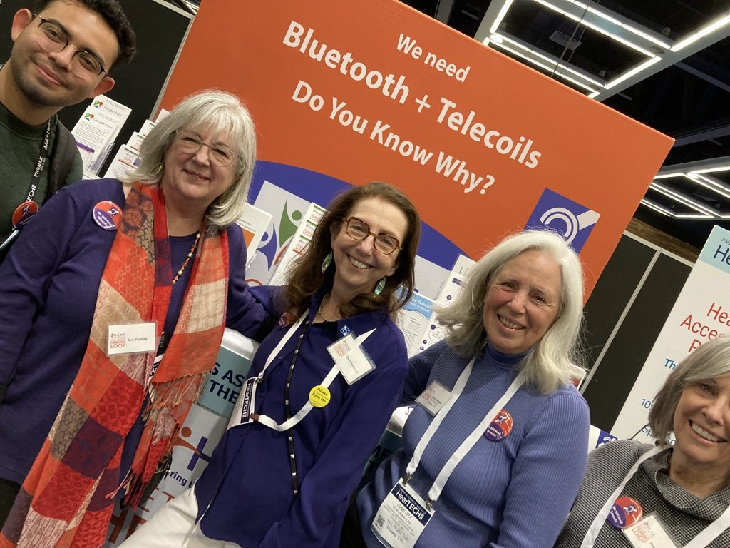 Image shows 4 hearing loss at the 2023 Annual Academy of Audiology Convention in Seattle in front of a large banner with the wording We need Bluetooth + Telecoils. Do you know why? 